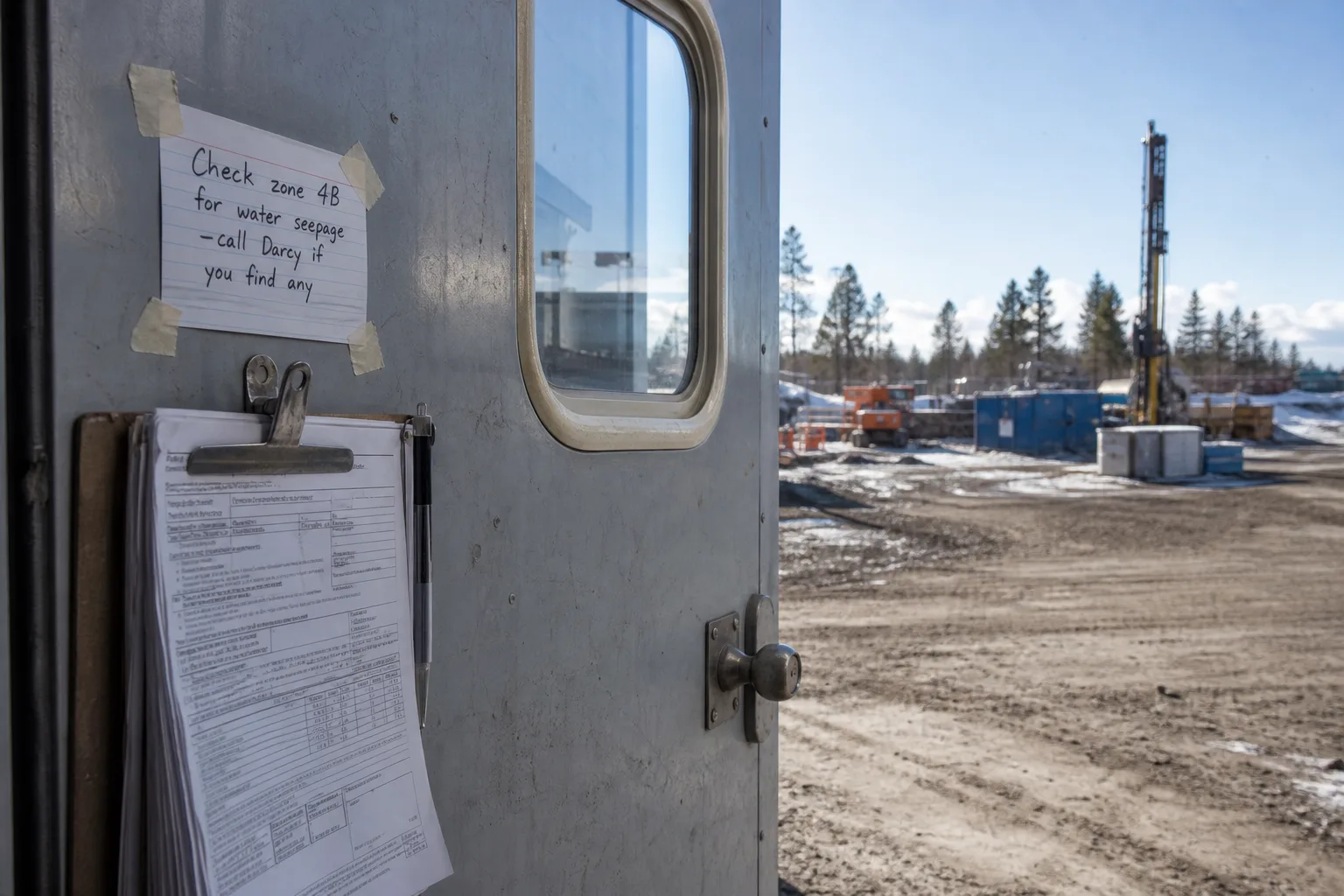 An index card with handwriting taped to the inside of a metal job trailer door, next to a clipboard of permit paperwork